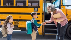 A young boy politely shakes hands with the bus driver while standing next to a school bus.