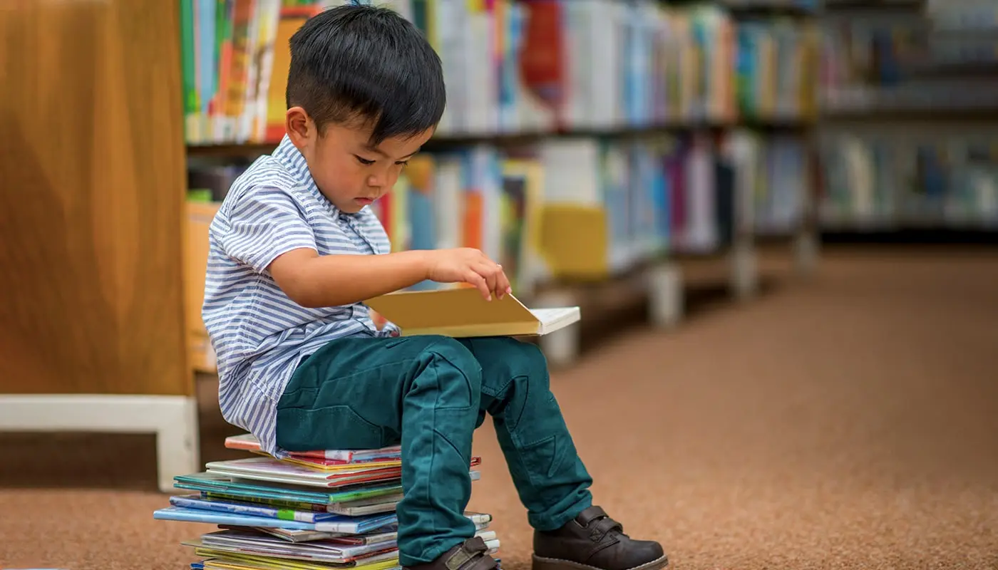 Reading is a big part of a child's growing up story. A boy sits on a stack of book while looking at another book.