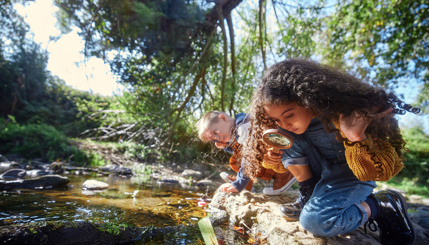 A young girl looks into a creek with a magnifying glass