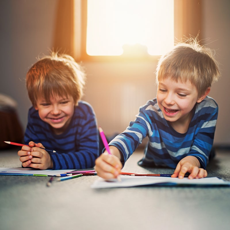 Two happy little boys are laying on the floor coloring.