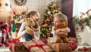 Two young girls laugh and hug bears in front of a Christmas tree on Christmas morning.