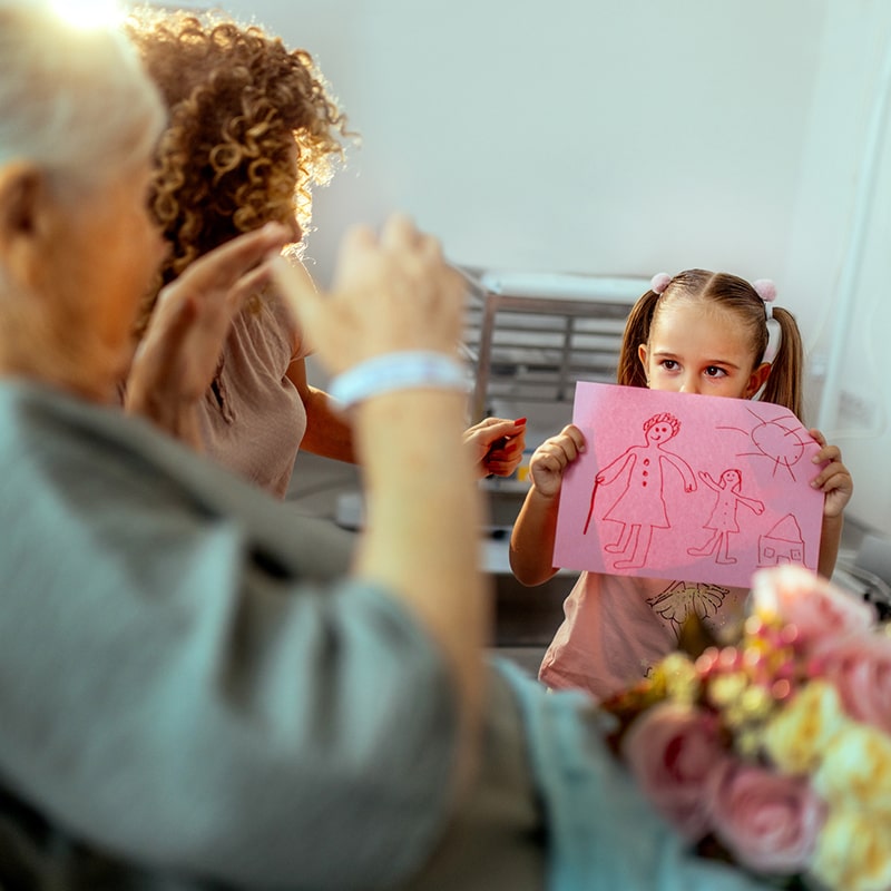 A young girl holds up a cute drawing she made and shows it to her grandmother who is sick in the hospital.