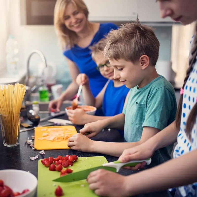 A happy mom looks on as her two young sons and older daughter cut up items for a build your own pasta bowl bar.