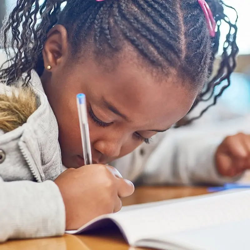 A focused young girl practices writing her name in a notebook while sitting at a desk.