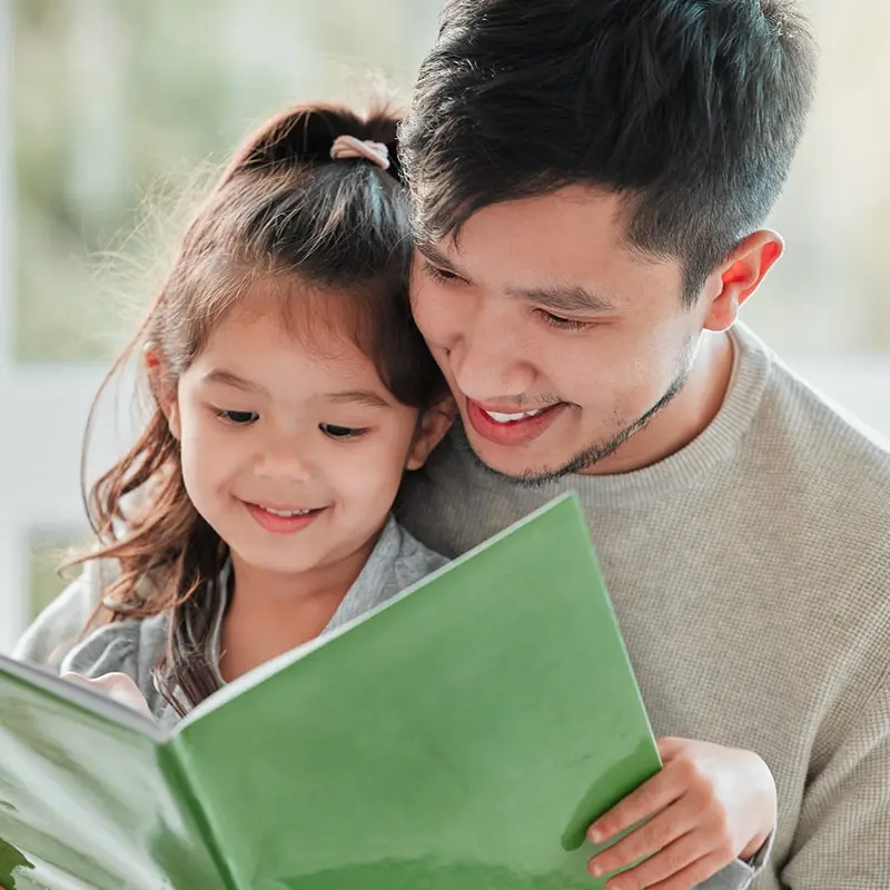 A smiling young girl sits in her father’s lap while they read a book together.